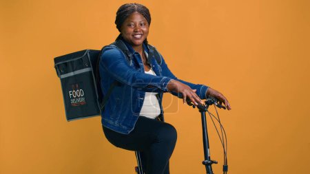 Téléchargez les photos : Courrier afro-américain fiable avec sac à l'arrière livrant de la nourriture du restaurant à un quartier local. Femme noire en bonne santé utilisant le vélo comme mode de transport pour une livraison efficace. - en image libre de droit