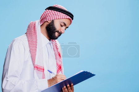 Photo for Man wearing traditional islamic thobe and headscarf clothes writing in clipboard, filling application form. Arab dressed in checkered ghutra and robe taking notes in checklist while posing in studio - Royalty Free Image