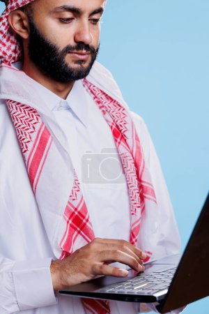 Téléchargez les photos : Homme vêtu de thobe musulman traditionnel et utilisant un pavé tactile pour ordinateur portable en gros plan. Arabe portant des vêtements islamiques, tenant un ordinateur portable et faisant défiler la page tout en naviguant sur Internet - en image libre de droit
