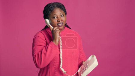 Téléchargez les photos : Femme afro-américaine en tenue élégante ayant une conversation par téléphone combiné. Blogueuse de mode féminine parlant sur un téléphone fixe devant un fond rose isolé. - en image libre de droit