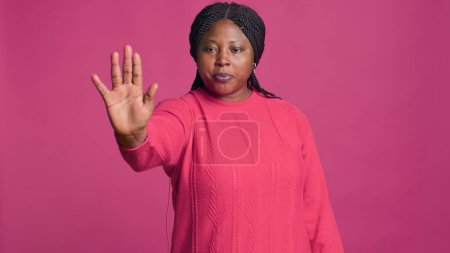 African american lady with denying using stop hand gesture towards camera against isolated background. Female fashion influencer disapproving rejecting disagree portrait on pink background.