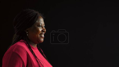 Foto de Mujer afroamericana confiada posa para un retrato encantador con un suéter rosa brillante. Vista lateral de la joven mujer negra con ropa de moda sonriendo a la cámara para una fotografía. - Imagen libre de derechos
