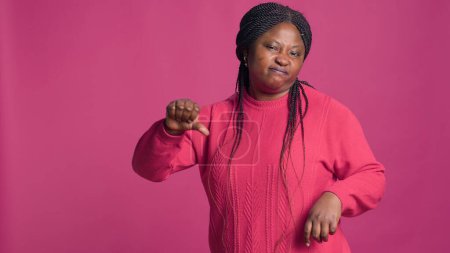 Téléchargez les photos : Expressive femme afro-américaine en pull rose gesticulant les pouces vers la caméra. Modèle de mode affichant désapprobation avec ses mains en face de fond isolé audacieux. - en image libre de droit