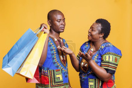 Photo for Smiling african american woman enjoying shopping spree while man carrying heavy paper bags. Cheerful wife happy with purchase while bored husband holding paperbags on shoulder - Royalty Free Image