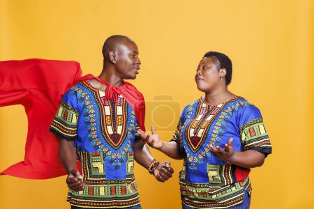 Photo for African american woman gesticulating and looking at man dressed in superman cape. Smiling boyfriend wearing red superhero cloak having communication with girlfriend in studio - Royalty Free Image