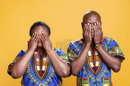 Photo for African american couple covering eyes with arms, showing hear no evil three wise monkeys portrait. Black man and woman pair holding palms on face on orange studio background - Royalty Free Image