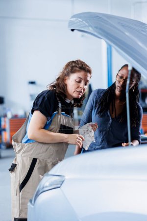Photo for Engineer in garage finishing fixing car for BIPOC woman, looking underneath vehicle hood to remove remaining oil leaks. Worker does routine exhaust system cleaning on client automobile - Royalty Free Image