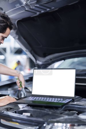 Photo for Mockup laptop on car with hood open while mechanic in blurry background replaces compressor belt. Isolated screen device next to skilled garage worker servicing client vehicle - Royalty Free Image