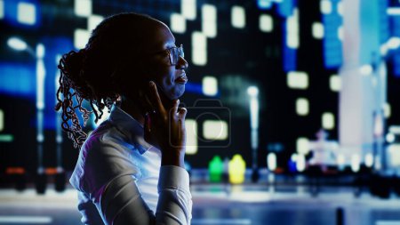 Photo for African american woman talking over the phone with husband using cellphone to feel more safe strolling around dimly illuminated empty avenues at night, blurry urban background with bokeh lights - Royalty Free Image
