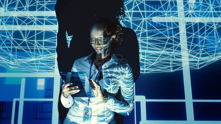 Foto de Mujer de apoyo tecnológico afroamericano que mira holograma de simulación cerebral humana de inteligencia artificial. Gerente de supervisión utiliza la tableta para aprovechar la energía de la tecnología AI, interactuando con la visualización de AR - Imagen libre de derechos
