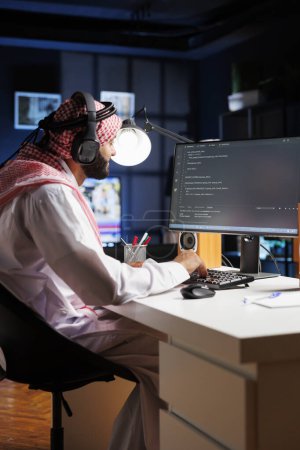 Photo for Data developer from the Middle East types code on a computer, demonstrating the core of software programming and development. Muslim man using wireless headphones and immersed in algorithm execution. - Royalty Free Image
