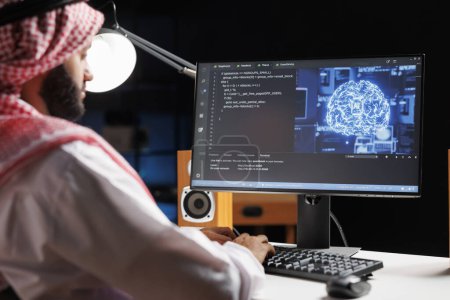 Photo for Muslim engineer focuses on a computer screen displaying code and data in a futuristic work setting. He manages a robust cloud-based system while monitoring data on a desktop computer screen. - Royalty Free Image