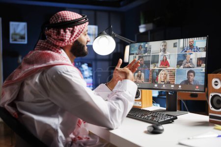 Téléchargez les photos : Image montrant un homme du Moyen-Orient portant des vêtements traditionnels, parlant avec un groupe de personnes sur son écran d'ordinateur. Un homme islamique conversant avec son équipe lors d'une conférence téléphonique. - en image libre de droit