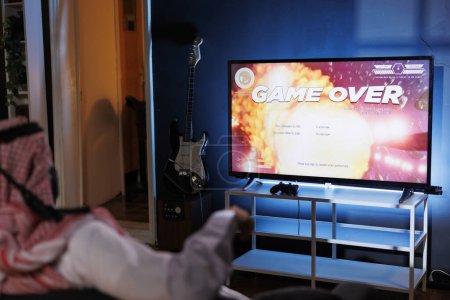 Photo for Islamic guy defeated in an online video game from the comfort of his home. Lit by the glow of the screen, he grasps a controller and uses a wireless joystick for immersive gameplay. - Royalty Free Image