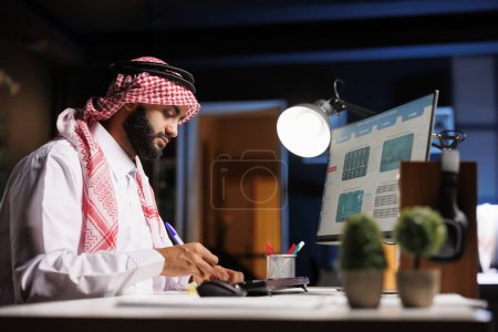 Foto de Un hombre de negocios musulmán con atuendo tradicional usa un bolígrafo para anotar notas de la pantalla de su computadora. La imagen retrata a un tipo árabe diligente que lleva a cabo un estudio de investigación con una PC de escritorio y bloc de notas. - Imagen libre de derechos