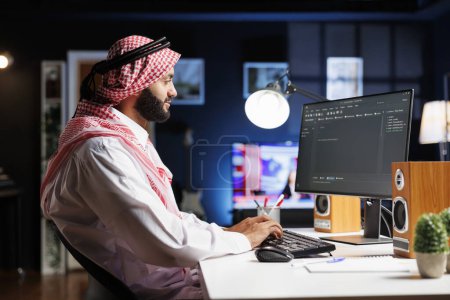 Téléchargez les photos : L'image montre un développeur arabe travaillant sur le moniteur PC, assis à un algorithme de traitement de poste de travail. Muslim Coder utilise un ordinateur de bureau pour travailler sur l'interface utilisateur. - en image libre de droit