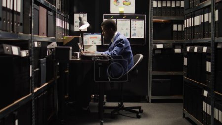 Téléchargez les photos : Un détective privé afro-américain entre dans la salle d'archives avec un sac d'ordinateur portable et commence une enquête criminelle. Homme enquêteur assis au bureau avec ordinateur sans fil, la vérification des dossiers suspects. - en image libre de droit
