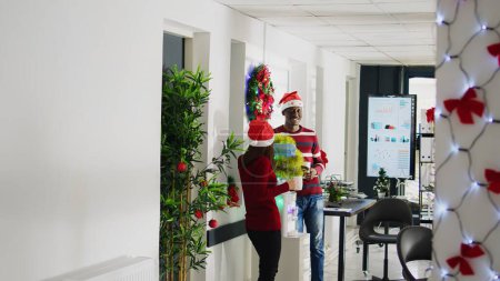 Téléchargez les photos : Les employés dans le bureau décoré de Noël profitant d'une tasse de café près de la glacière à eau, riant ensemble. Heureux membres du personnel du BIPOC portant des chapeaux du Père Noël prenant une pause dans un espace de travail multiracial - en image libre de droit
