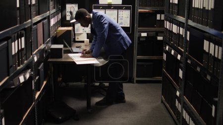 Photo for African American policeman carefully analyzes material, does records searches, and collects data from shelves and laptop. Male police officer standing, looking through case files on desk in office. - Royalty Free Image