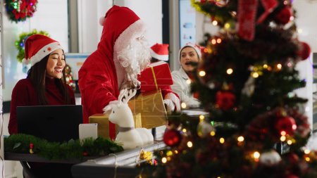 Foto de Los trabajadores multiétnicos que reciben regalos de un colega que finge ser Santa Claus en la oficina condecorada con Navidad. Empleado con traje sorprendente personal de la compañía con regalos durante la temporada de vacaciones de invierno - Imagen libre de derechos