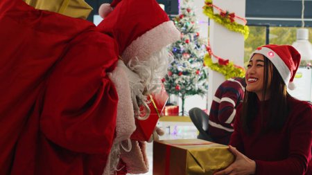 Foto de Primer plano del gerente vestido como Santa Claus difundiendo alegría navideña en la oficina de adornos festivos, ofreciendo regalos a los empleados. Supervisor sorprendente emocionados trabajadores de la empresa con regalos de Navidad - Imagen libre de derechos