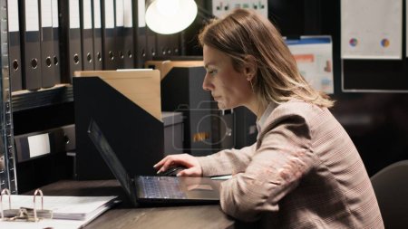 Photo for Policewoman captured in midst of investigation, examining records and evidence. Detailed view of officer at desk with laptop and case files opened for cross-referencing suspect profile and statements. - Royalty Free Image