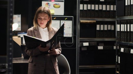 Female private detective conducting investigation in office, focused on reviewing files and evidence. Portrait shot of policewoman analyzing statements and records. Looking at camera, tripod shot.