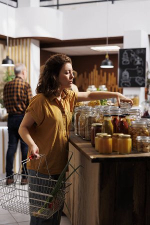 Photo for Woman holding shopping basket, buying ecofriendly bulk products in zero waste store designed to minimize plastic usage. Green living customer looking for nutritious groceries - Royalty Free Image