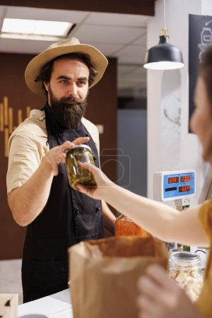 Photo for Woman at checkout counter in zero waste shop, buying locally sourced ecofriendly vegetables from happy storekeeper. Client purchasing pickles jar in local neighborhood store using renewable energy - Royalty Free Image