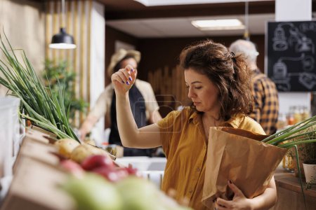 Foto de Mujer en el mercado de agricultores comprando verduras libres de químicos, recogiendo cebollas verdes maduras. Cliente en una tienda de comestibles local libre de plástico usando una bolsa de papel desmontable para evitar desperdicios excesivos - Imagen libre de derechos