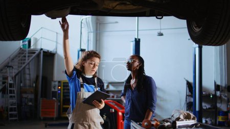 Photo for Mechanic and customer standing underneath car in garage using laptop to order new parts after finding fire hazards. Expert assisting client by looking for components online, zoom out shot - Royalty Free Image