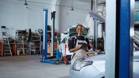 Photo for Portrait of cheerful engineer working in professional garage, using tablet to assess car performance before making necessary upgrades. Joyful repair shop worker tuning up vehicle - Royalty Free Image