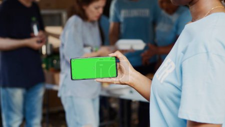 Photo for Black woman holds smartphone with isolated chromakey template for customized charitable messages. African American person at an outdoor food drive with a cellphone displaying a green screen. - Royalty Free Image