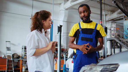 Photo for Experienced serviceman in car service showing customer what needs to be changed on her automobile for it to properly work. Garage professional inspecting vehicle placed on lift, chatting with client - Royalty Free Image
