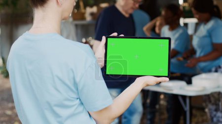 Photo for Caucasian woman horizontally holding digital tablet with green screen chromakey display at an outdoor food bank. Smart device displaying blank mockup template held by female charity worker, outdoors. - Royalty Free Image