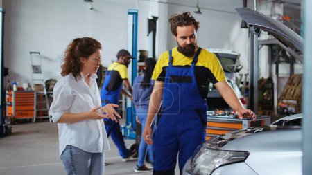Photo for Adept mechanic helping client with car maintenance in auto repair shop. Employee in garage facility looking over automobile parts with woman, mending her vehicle during yearly inspection - Royalty Free Image