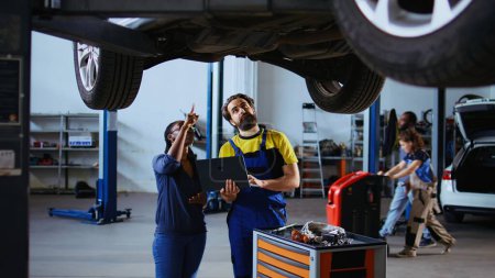 Photo for Serviceman and customer standing underneath car in garage using laptop to order new parts after finding damages. Employee assisting client by looking online for replacing automobile components - Royalty Free Image