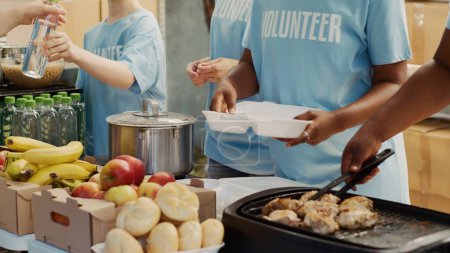 Téléchargez les photos : Des personnes multiethniques en t-shirts bleus, rassemblées à l'extérieur pour distribuer des dons de nourriture et aider les sans-abri et les affamés. Mettre l'accent sur les bénévoles qui servent gratuitement des repas fraîchement préparés aux personnes dans le besoin. - en image libre de droit