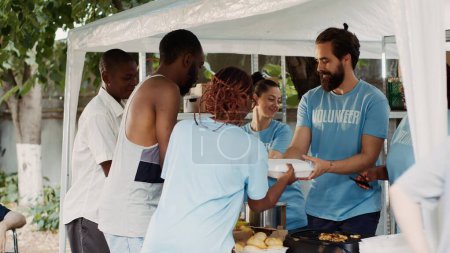 Foto de La organización caritativa multiétnica proporciona comida y provisiones gratuitas a las personas sin hogar y lisiadas. Mujer negra vistiendo una camiseta azul ayudando a un chico afroamericano indefenso, con muletas. - Imagen libre de derechos