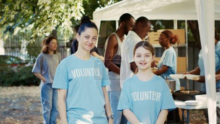 Photo for Portrait shot of mother and daughter participating in hunger relief program at outdoor food bank. Caucasian volunteers in blue t-shirts and looking at camera, ready to assist the needy. Tripod shot. - Royalty Free Image