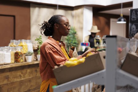 Téléchargez les photos : Une cliente afro-américaine dans une épicerie. Elle parcourt les légumes biologiques, les céréales et les produits durables. Le magasin propose des produits locaux zéro déchet. Mise au point. - en image libre de droit