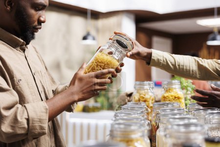 Téléchargez les photos : Gros plan d'un Afro-Américain regardant les pâtes dans les pots de verre. Dans un supermarché soucieux de l'environnement, un client masculin à la recherche de produits en vrac nutritifs. - en image libre de droit