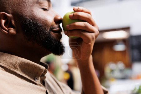 Photo for Selective focus on black man at zero waste store smelling an organic farm-grown apple. Close-up of African American male customer trying to determine if local neighborhood shop fruits are fresh. - Royalty Free Image