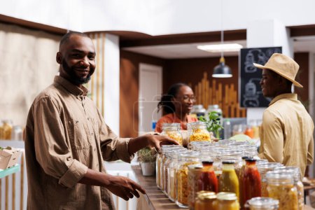 Photo for A smiling African American man looking at the camera in a modern grocery store. Reusable packaging, eco friendly products, and a variety of items are showcased on the shelves. - Royalty Free Image