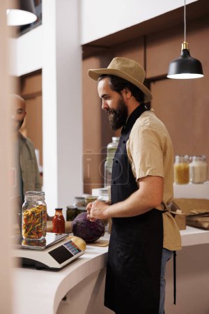 Photo for Male storekeeper, wearing a hat and apron, stands behind cashier desk while looking at jar filled with certain organic food item on measuring scale. Caucasian vendor weighing pasta for customer. - Royalty Free Image