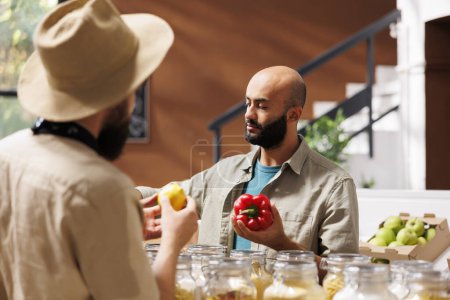Téléchargez les photos : Un client du Moyen-Orient tient un poivron rouge tandis qu'un commerçant le regarde tout en tenant un citron, dans une épicerie respectueuse de l'environnement. Vendeur conversant avec le client sur un mode de vie sain. - en image libre de droit