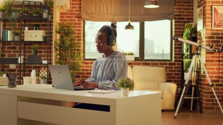 Photo for Freelancer enjoys working at workstation while listening to music and performing internet activities. At sunset, african american woman is multitasking, online career in blogging. - Royalty Free Image