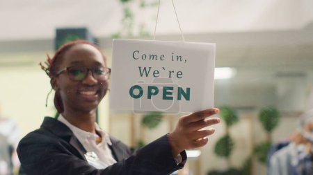 Foto de Empleado afroamericano abriendo tienda de ropa por la mañana, esperando clientes. Cartel de mensaje cerrado en la puerta boutique de moda en el centro comercial - Imagen libre de derechos
