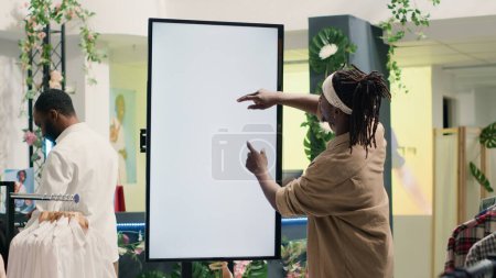 Foto de Hombre afroamericano en la tienda de ropa usando la pantalla de maqueta de realidad aumentada para ver las opciones de ropa para probarse. Cliente que utiliza la pantalla led para visualizar las opciones de moda en la sala de exposición - Imagen libre de derechos