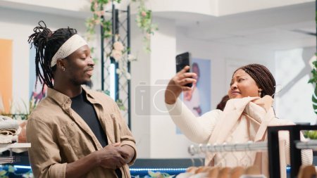 Photo for Man in fancy fashion boutique convincing his girlfriend to buy stylish formal clothes on hangers. Customers in premium showroom debating whether to purchase new designer clothing collection or not - Royalty Free Image